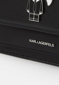 Musta nahkainen asuste, jossa on ommeltu reuna, metallinen Karl Lagerfeld -logo ja pieni hopeinen hahmo puvussa ja solmiossa.