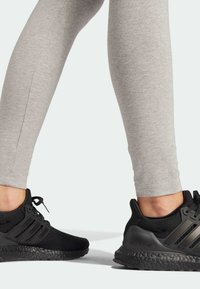 Fekete sportcipők hálós felső résszel és három fekete csíkkal, szürke testhez simuló leggingszel párosítva, amely felfedi a bokát és a texturális részleteket.
