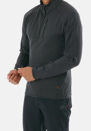 Mann trägt dunkelgrauen langärmeligen Pullover mit Reißverschluss und Brusttasche und schwarze Hose, die Hände vor der Taille verschränkt.