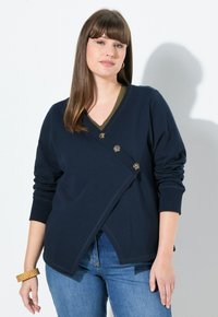 Cardigan avvolgente blu navy con scollo a V, dotato di tre bottoni marroni. Ha maniche lunghe e una finitura testurizzata, abbinato a jeans blu.