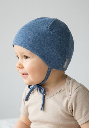 Baby trägt eine blaue gestrickte Mütze, die unter dem Kinn gebunden ist, lächelt und schaut nach links, gekleidet in ein beiges Kurzarmhemd.