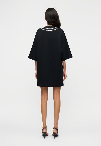 Μαύρο φαρδύ κοντομάνικο φόρεμα με κοντά μανίκια, τονισμένο με λευκή λεπτομέρεια σε σχήμα μαργαριταριού γύρω από τον λαιμό, με απαλή υφή υφάσματος.