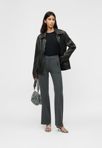 Γυναίκα που φοράει σκούρο δερμάτινο μπουφάν, μαύρο πουκάμισο, γκρι παντελόνι με ραφή, ψηλοτάκουνα πέδιλα, κρατώντας μια τσάντα με σχέδιο σε λευκό φόντο.