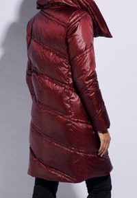 Manteau rembourré bordeaux avec capuche, présentant un design matelassé, une texture brillante et des coutures en diagonale. Coupe ajustée avec des manches longues.