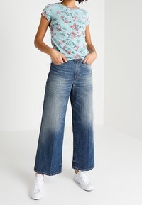 Camiseta floral de color azul claro con mangas cortas, combinada con jeans de pierna ancha de un azul desgastado y zapatillas blancas.