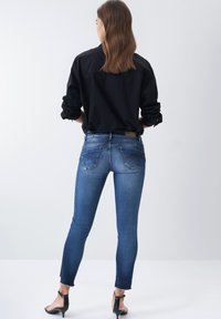 Vaqueros ajustados en denim azul medio con un dobladillo deshilachado, bolsillos traseros acentuados y un acabado suave. Combinados con una camisa de botones negra.
