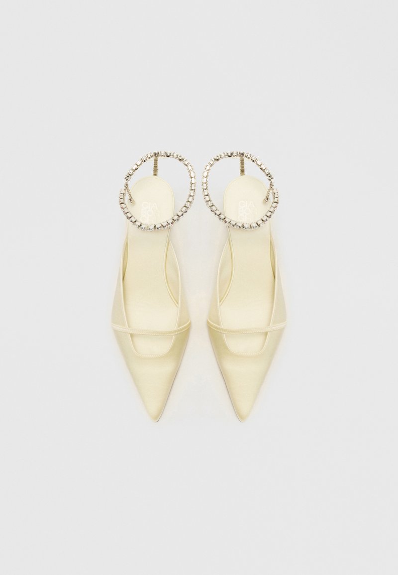 Zapatos de tacón beige de satén con punta afilada, que presentan una correa transparente y adornos de strass en forma circular en el tobillo. Suela plana.