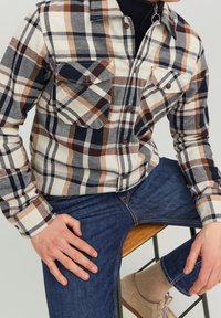 Camicia a scacchi in crema, blu navy, marrone e arancione con due tasche sul petto, chiusura con bottoni e maniche lunghe, abbinata a jeans in denim e scarpe.