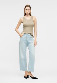 Γυναίκα που φοράει μπεζ αμάνικη πλεκτή μπλούζα, ανοιχτό μπλε τζιν παντελόνι με φαρδιά μπατζάκια και ξεφτισμένα τελειώματα, και μαύρα παπούτσια με μυτερή μύτη, στέκεται με το ένα χέρι στην τσέπη.