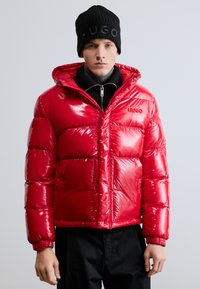 Piros, fényes pufi dzseki kapucnival, cipzáras záródással és elasztikus mandzsettákkal. Fekete garbó fölött viselve, fekete kötött sapkával párosítva.