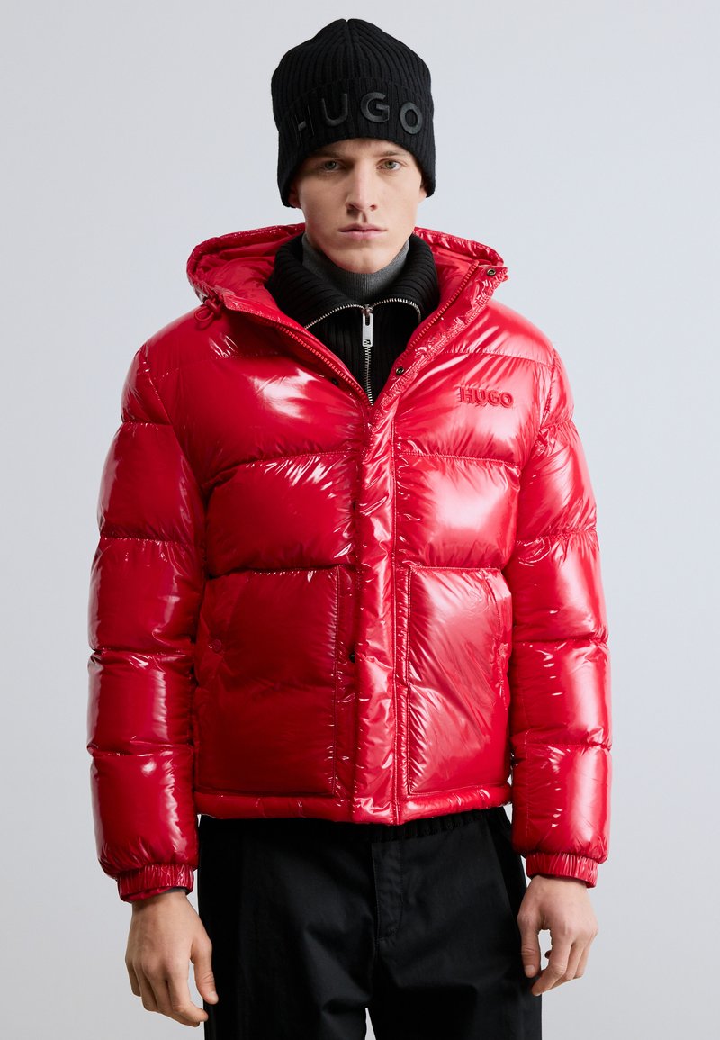 Piros, fényes pufi dzseki kapucnival, cipzáras záródással és elasztikus mandzsettákkal. Fekete garbó fölött viselve, fekete kötött sapkával párosítva.