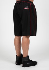 Zwarte shorts met rode zijstrepen, met een logo aan de linkerzijde en zakken. De structuur lijkt glad en de stof is lichtgewicht.