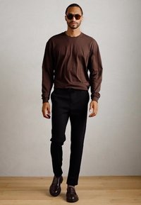 Hombre que lleva una camisa larga marrón, pantalones negros, zapatos de cuero marrones y gafas de sol, caminando sobre un suelo de madera clara frente a una pared gris.