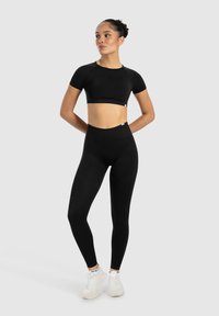 Schwarzes Sportset mit einem figurbetonten Crop-Top und hochgeschnittenen Leggings, beide aus glattem, dehnbarem Stoff, kombiniert mit weißen Sportschuhen.