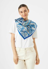 Écharpe en soie ornée d'un motif paisley bleu avec des accents en teal et or, drapée autour du cou sur une chemise blanche.