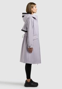 Femme vêtue d'un imperméable à capuche gris clair avec une taille à cordon, leggings noirs et baskets noires, debout de profil sur un fond neutre.