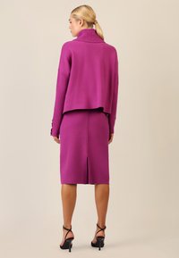Conjunto de punto fucsia que incluye un suéter corto de canalé con cuello alto y una falda lápiz con una abertura trasera, combinado con tacones negros de tiras.