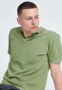 Polo vert à manches courtes avec un col en V, tissu texturé et broderie subtile du logo sur la poitrine. Coupe décontractée, texture lisse.