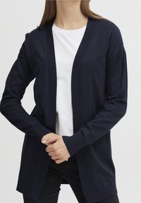 Lång marinblå cardigan i stickad mjukvara, med en öppen framdesign, ribbade manschetter och en avslappnad passform. Buren över en vit skjorta.