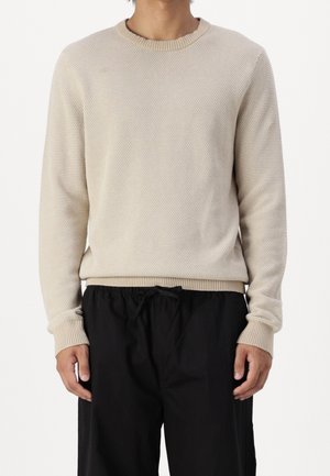 Beige stickad tröja med en texturerad mönster, rund halsringning och ribbad manschett, i kombination med svarta byxor med dragsko.