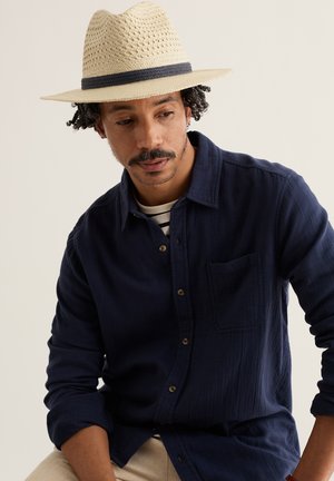 Man with short curly hair and mustache wearing a beige straw hat and dark blue button-up shirt, looking downward against a plain background.