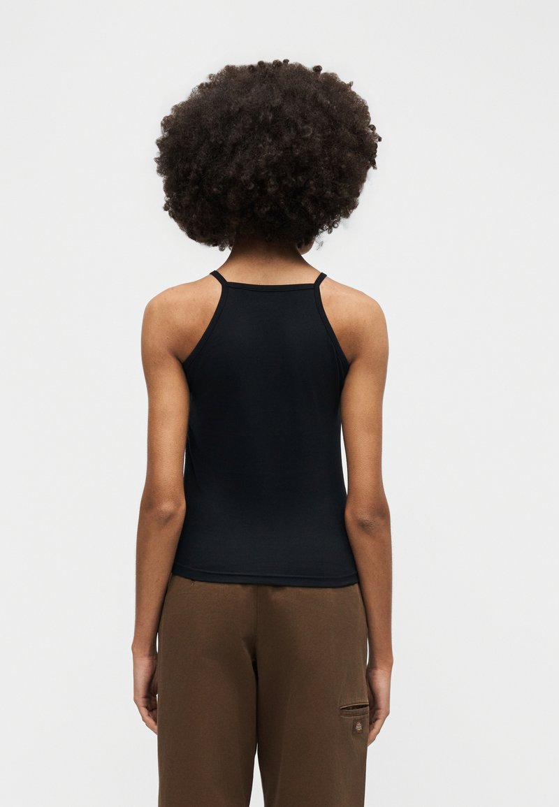 Person with curly hair wearing a black sleeveless top and brown pants, standing facing a plain light background.