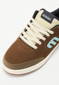 Bruna skate skor med ovandel i mocka, krämfärgade snören, en ljusblå logotyp och gummisula. Har en vadderad krage och förstärkt tå.