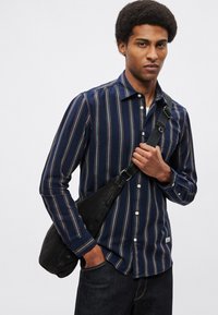 Granatowa koszula na guziki w pionowe czerwono-białe paski. Posiada kieszeń, długie rękawy oraz luźny krój, zestawiona z czarną torbą na ramię.