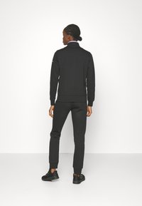 Sudadera negra y joggers a juego en un material suave y texturizado. Presenta un cuello alto y puños de canalé, combinados con zapatillas deportivas negras.