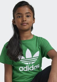 Zelené bavlněné tričko se širokým bílým logem trefoil Adidas a nápisy, kulatým výstřihem, krátkým rukávem a hladkou texturou.