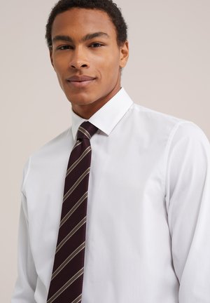 Un homme portant une chemise blanche impeccable et une cravate bordeaux à rayures diagonales se tient devant un fond clair uni.