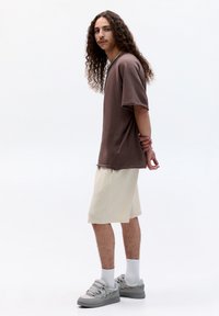 Braunes, übergroßes Kurzarm-T-Shirt, cremefarbene Shorts und graue Sneakers. Merkmale sind der lockere Sitz und eine subtile Textur, vor einem schlichten Hintergrund.