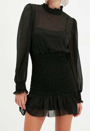 Femme portant une robe noire transparente à manches longues avec des épaules bouffantes, une taille smockée, des poignets à volants et un ourlet volanté.