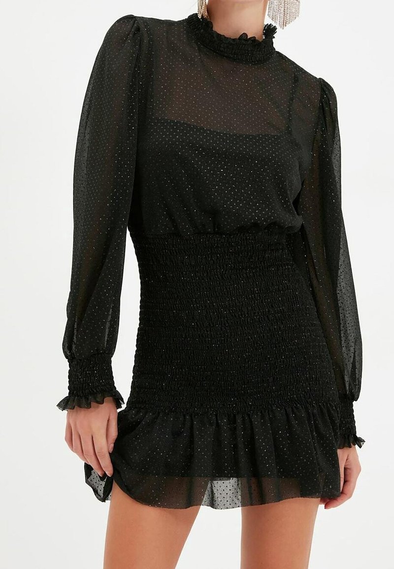 Femme portant une robe noire transparente à manches longues avec des épaules bouffantes, une taille smockée, des poignets à volants et un ourlet volanté.