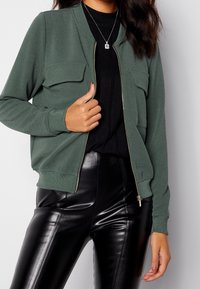 Giacca bomber verde con chiusura a zip, due tasche sul petto, polsini e vita a coste. Indossata sopra un top nero e abbinata a pantaloni neri lucidi.
