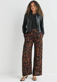 Vrouw met krullend haar, gekleed in een zwarte leren cropped jack, een zwart top, wijde bruine en zwarte panterprint broek en zwarte hakken.