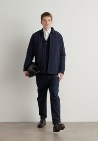Chaqueta de navy acolchada con dos bolsillos en el pecho, superpuesta sobre un suéter oscuro y una camisa blanca, combinada con pantalones oscuros y botas negras.