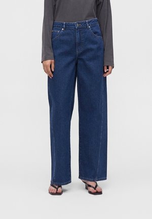 Personne portant un jean taille haute bleu foncé à jambes larges, un haut à manches longues foncé et des sandales à talons ouvertes noires sur un fond uni.