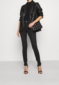 Blazer de cuero negro sobre una blusa negra acanalada, combinado con jeans oscuros y tacones altos negros con tiras. Un bolso negro acolchado completa el look.