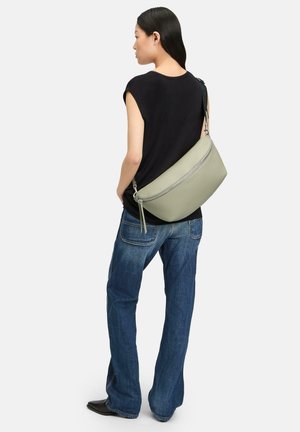 Oliwkowa skórzana torba na ramię o zaokrąglonym kształcie, z zamkiem błyskawicznym i detalem w postaci frędzli; w zestawie z prostą czarną bluzką i niebieskimi dżinsami.