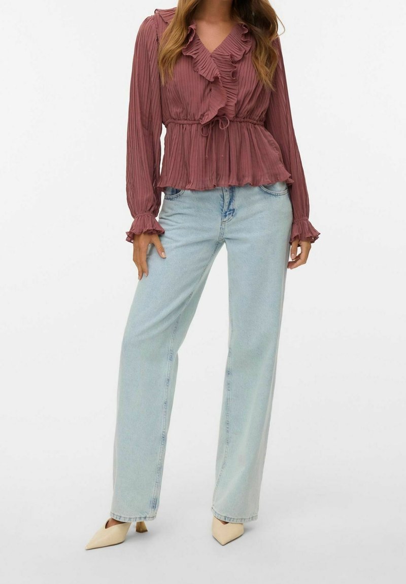 Femme portant un chemisier mauve à volants avec une ceinture à la taille, jean droit bleu clair, et escarpins beiges à bout pointu, debout sur un fond blanc.