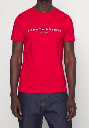 Mand iført en knald rød t-shirt med korte ærmer med "Tommy Hilfiger EST. 1985" logo og mørkeblå jeans mod en ensfarvet baggrund.