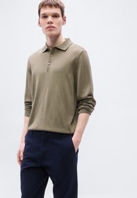 Πράσινο ελιάς πόλο με μακριά μανίκια, γιακά, δύο κουμπιά και κεντημένο λογότυπο. Συνδυάζεται με σκούρο μπλε παντελόνι, παρουσιάζοντας άνετη εφαρμογή.