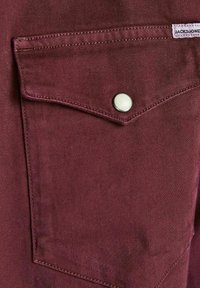 Gros plan d'une poche en tissu bordeaux avec rabat sécurisé par un bouton argenté et une étiquette en tissu blanche indiquant "JACK&JONES" sur le bord supérieur.