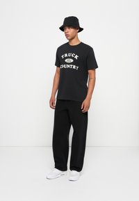 Juoda medvilninė marškinėliai su baltais užrašais „TRUCK“ ir „COUNTRY“; derinami su laisvai priglundančiomis juodomis kelnėmis ir baltais sportiniais bateliais. Juoda kibiro formos kepurė.