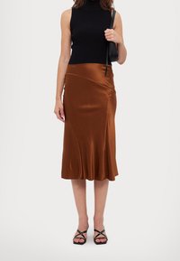 Falda de satén marrón con un dobladillo acampanado, combinada con una camiseta de tirantes negra de canalé y sandalias negras con tiras. Bolso sencillo sostenido en la mano.