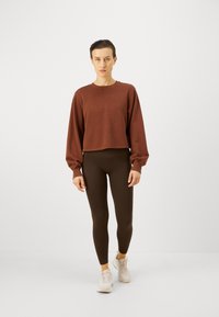 Brauner, taillierter Sweatshirt mit langen Ärmeln, kombiniert mit dunkelbraunen Leggings und hellen Turnschuhen. Angenehme Textur und schlichtes Design.