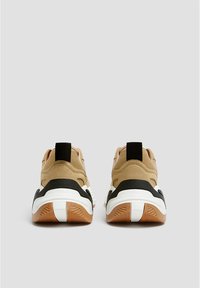 Baskets en daim tan avec une semelle noire et blanche, un dos texturé et une languette. Le design comprend des formes arrondies et des couleurs contrastées.