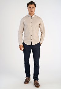 Camicia beige con bottoni, dal texture sottile, bottoni neri, abbinata a pantaloni blu scuro e scarpe da barca marroni con lacci rosa.