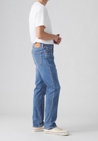 Homme debout en jean bleu et baskets blanches, mains jointes, portant un t-shirt blanc uni sur un fond gris clair.